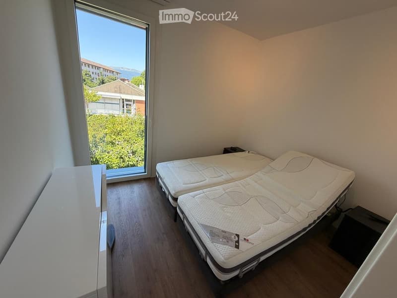 Modern, unfurnished bedroom with two single beds, wooden flooring, large window with view, and nightstand.