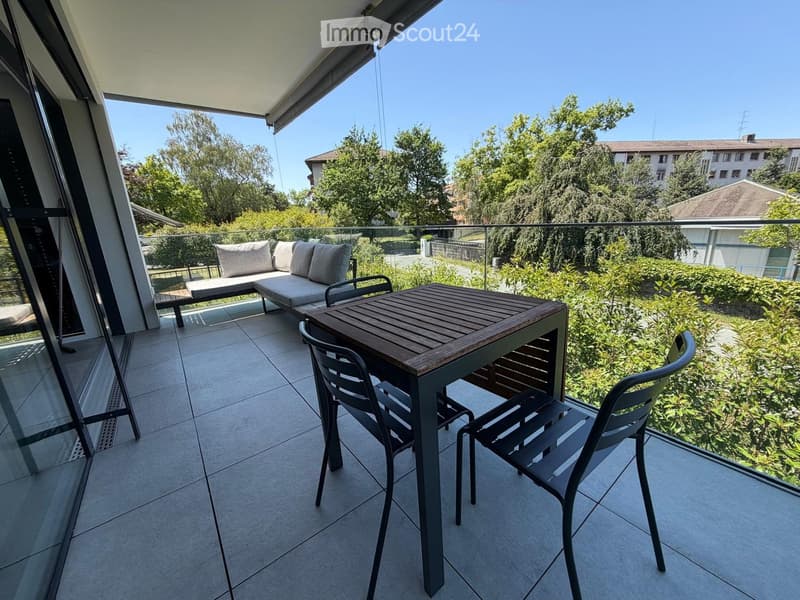Balcony with a sofa, a wooden table, and two chairs, overlooking a green area with trees and buildings.