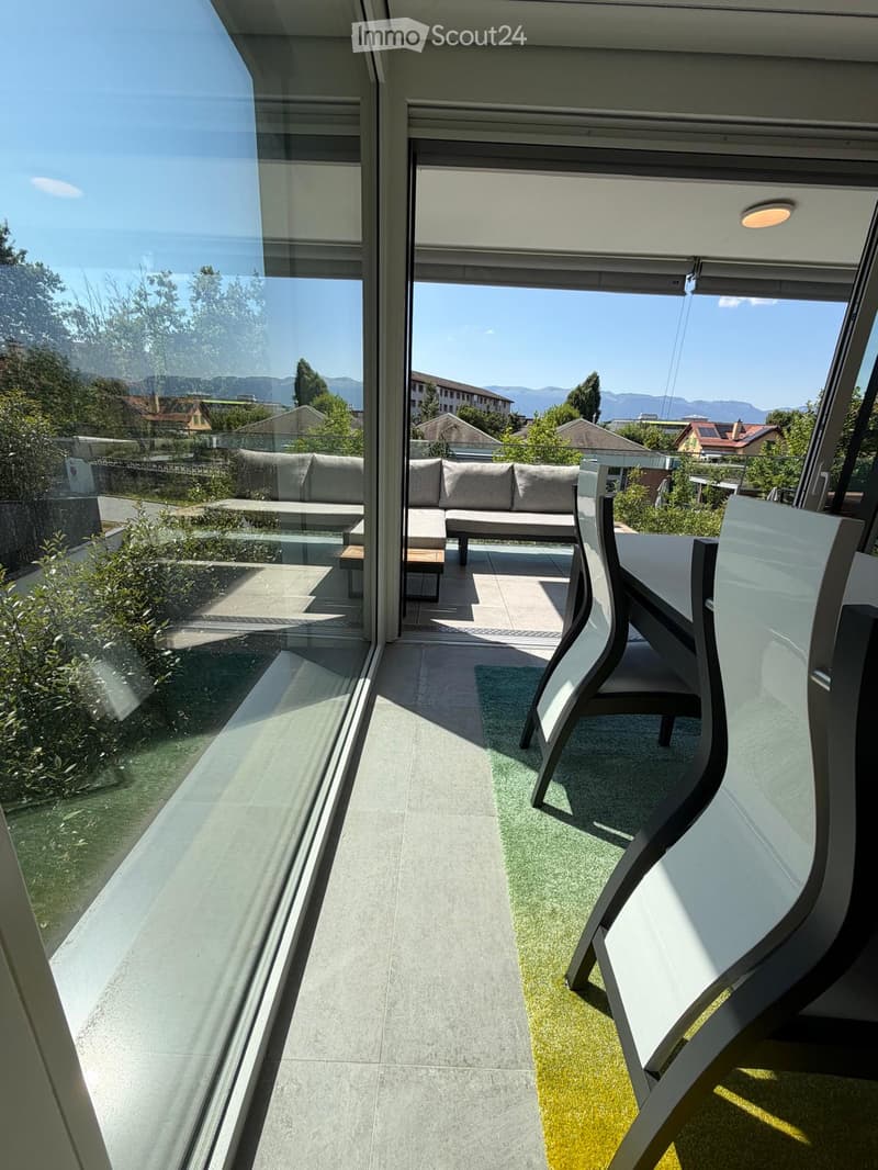 Modern balcony with large glass windows, grey sofa, white table, and chairs, overlooking a scenic view of trees and mountains.