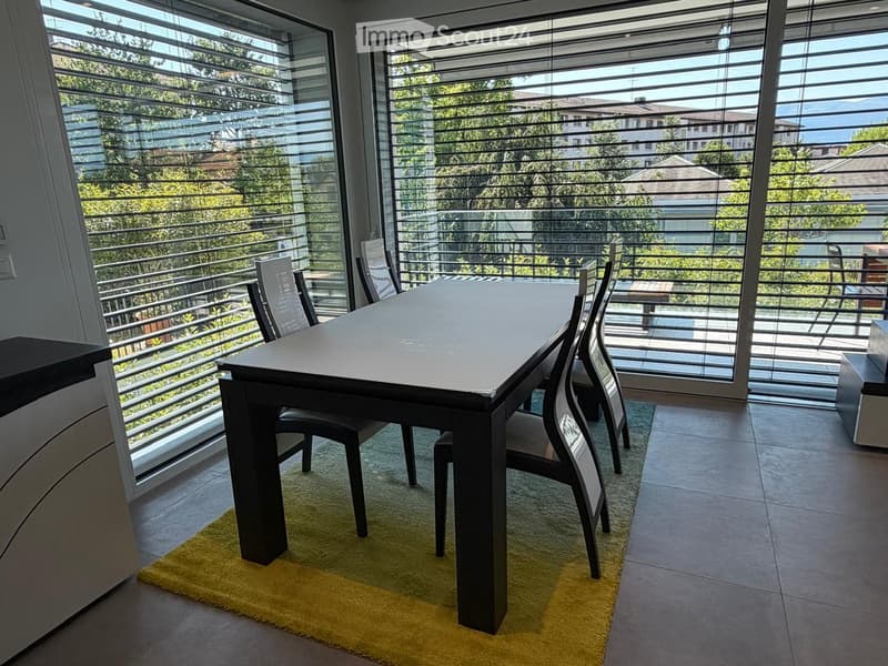 A modern dining area with a sleek black dining table and matching chairs, placed on a yellow rug. Large windows with blinds offer a scenic view of the surrounding greenery and buildings.