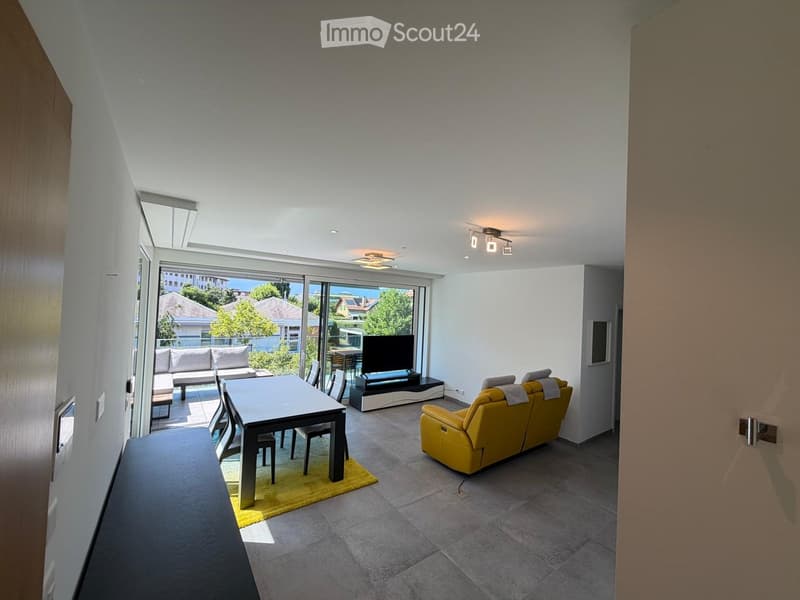 Modern living room with a yellow sofa, dining table, chairs, and a television. Large windows offer a view of the outdoor area.