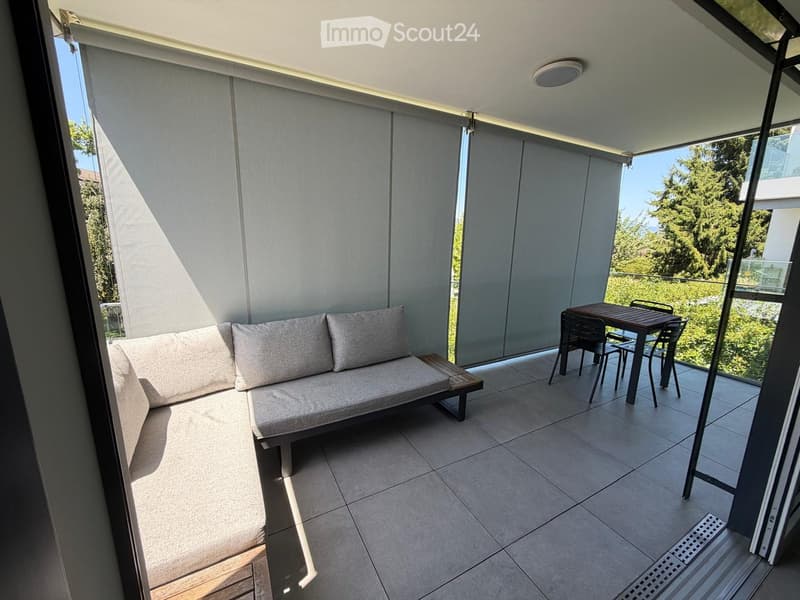 Modern balcony with tiled floor, grey sofa, small table, and chairs, large glass windows with blinds, and a view of greenery.