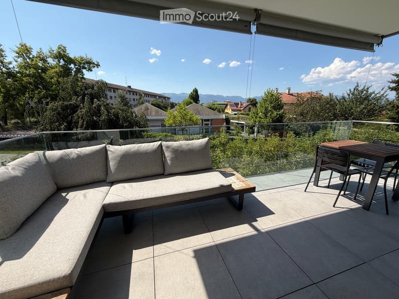 Balcony with a modern sofa, small table, and chairs, overlooking a green area with trees and buildings.