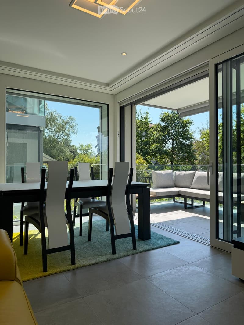 Modern dining area with a sleek black dining table, contemporary chairs, and a comfortable sofa visible through large glass doors leading to a balcony.