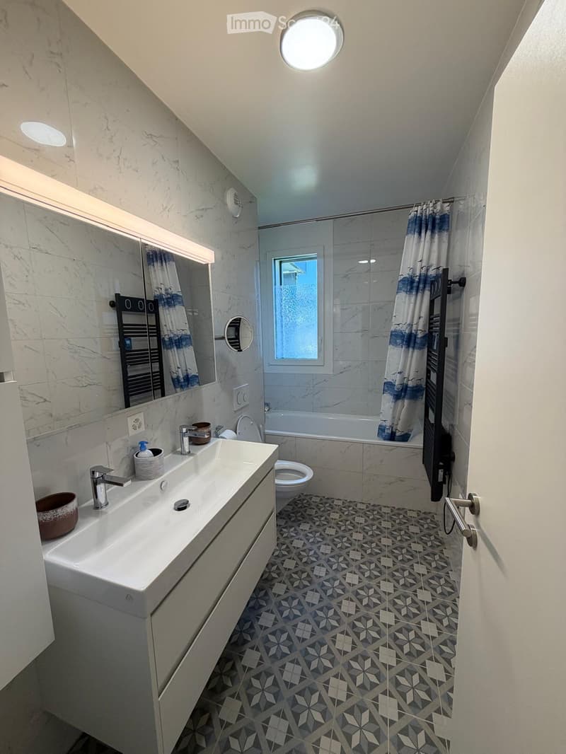 Modern bathroom with a white sink, chrome faucet, toilet, bathtub, and a large mirror. The floor features a geometric tile pattern, and the walls are clad in marble. A round skylight provides natural light.