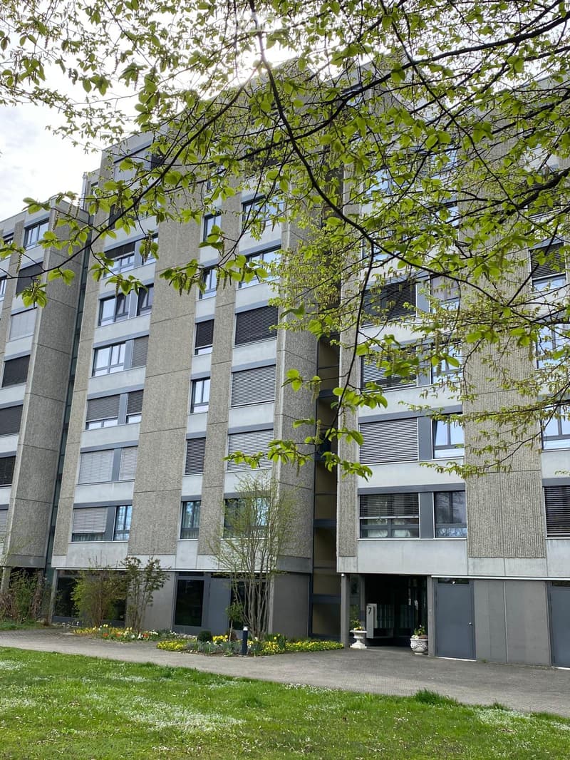 A multi-story residential building with gray exterior walls, multiple windows, and a well-maintained garden in front.