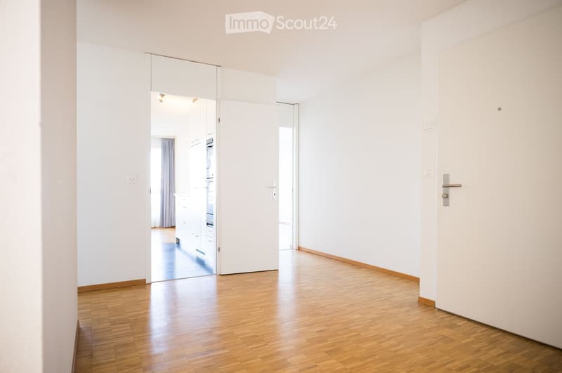 Empty room with wooden floor, white walls, open door, and a glimpse of a kitchen with a window and curtain.