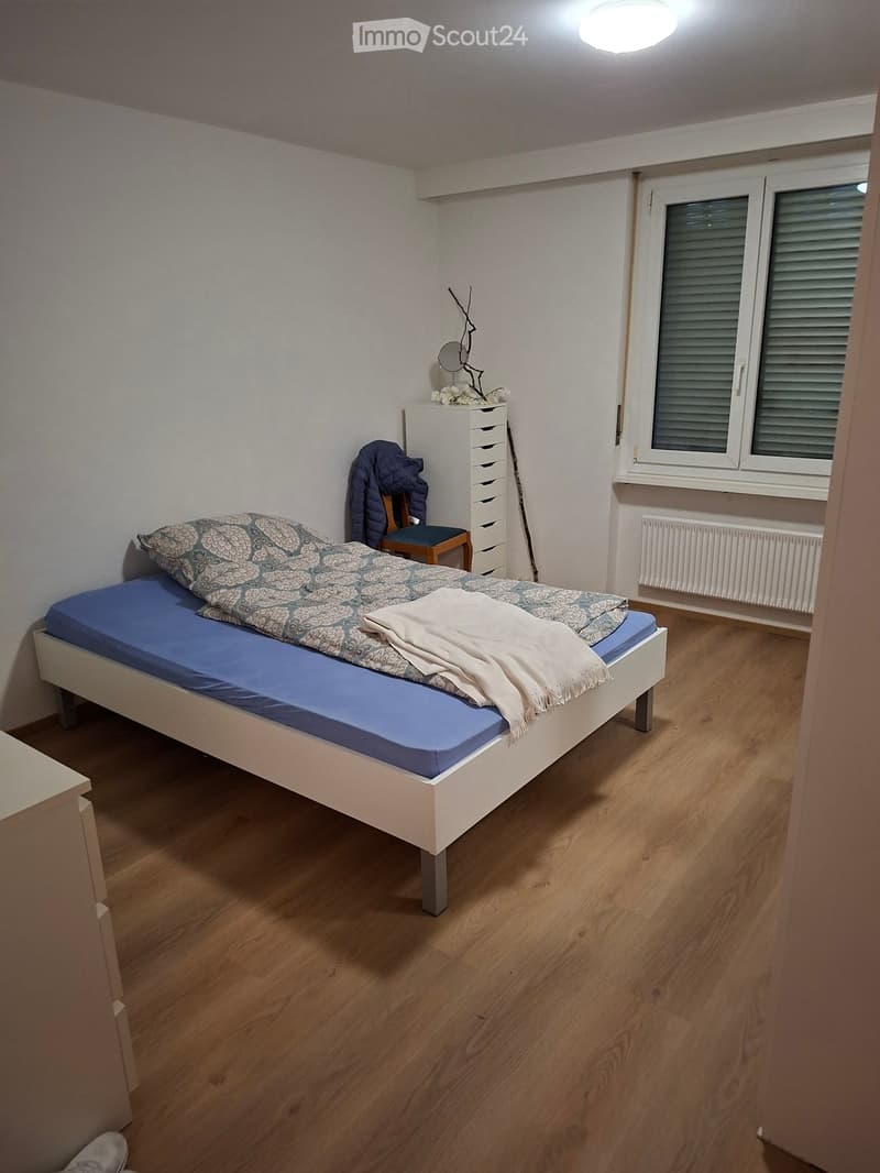 Cozy bedroom with a single bed, patterned bedding, a small chair with a jacket draped over it, a white drawer unit, and a mirror on the wall. The room has wooden flooring and a window with blinds.