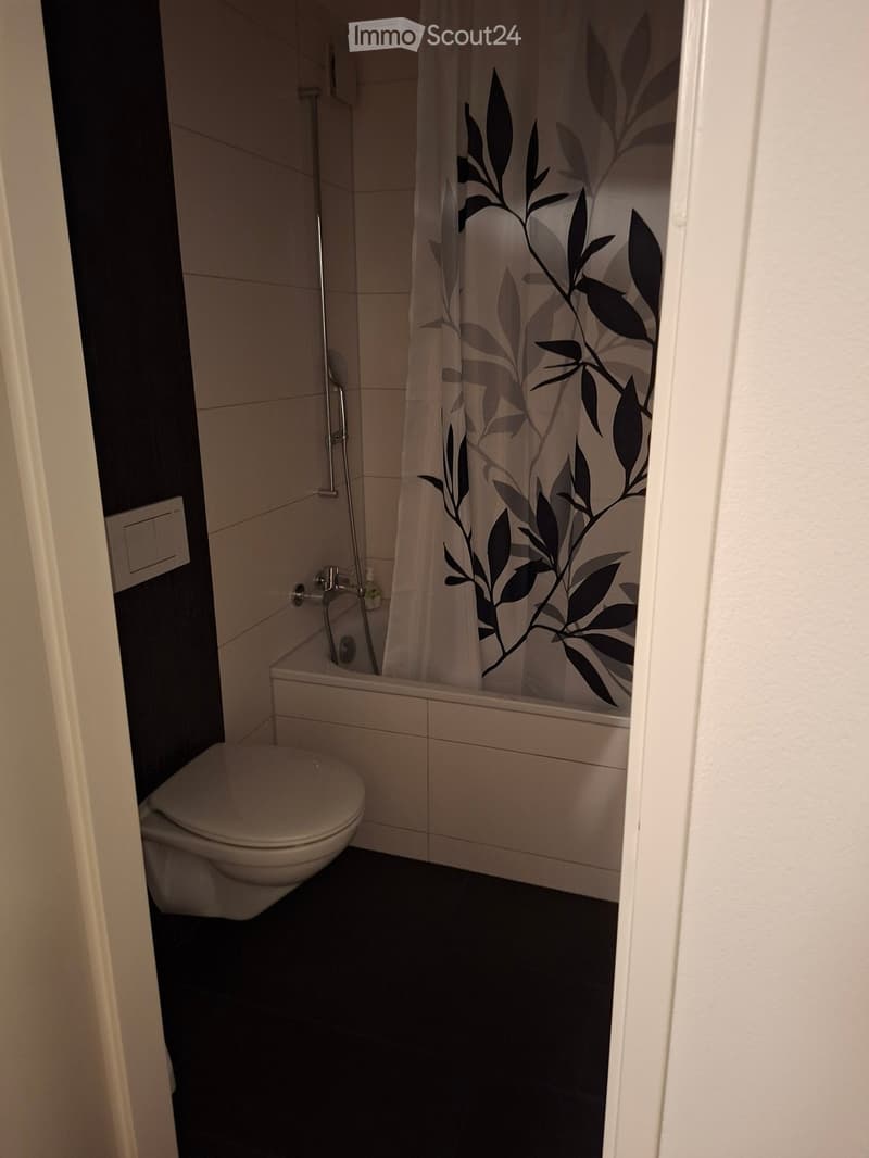 Modern bathroom with a toilet, bathtub, and a shower curtain featuring a leafy design. The walls are tiled, and the floor is dark.