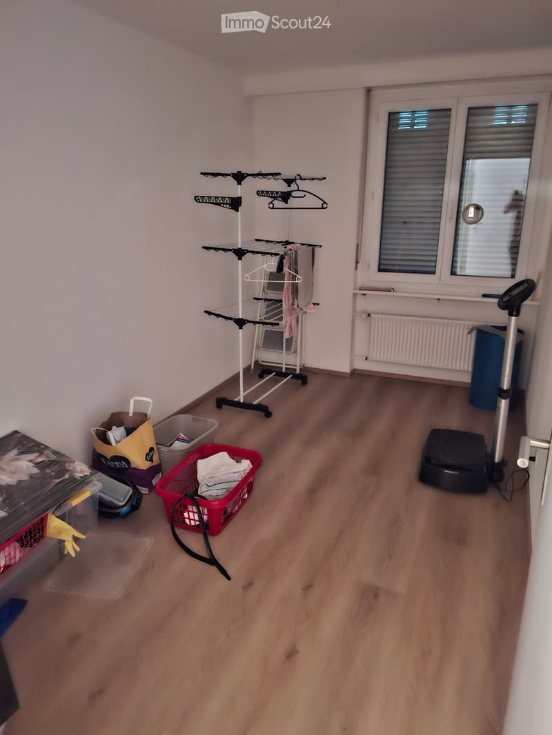 A small, unfurnished room with wooden flooring, a laundry rack, a shopping bag, a storage bin, an exercise bike, and some luggage.