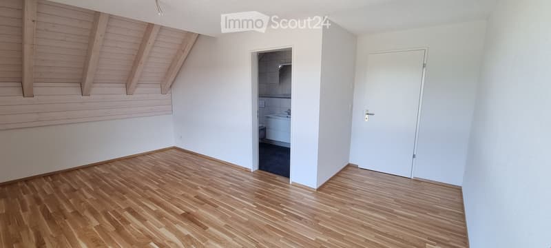 Empty room with wooden flooring, white walls, and exposed wooden beams on the ceiling. A white door leads to a small bathroom with a bathtub and toilet.