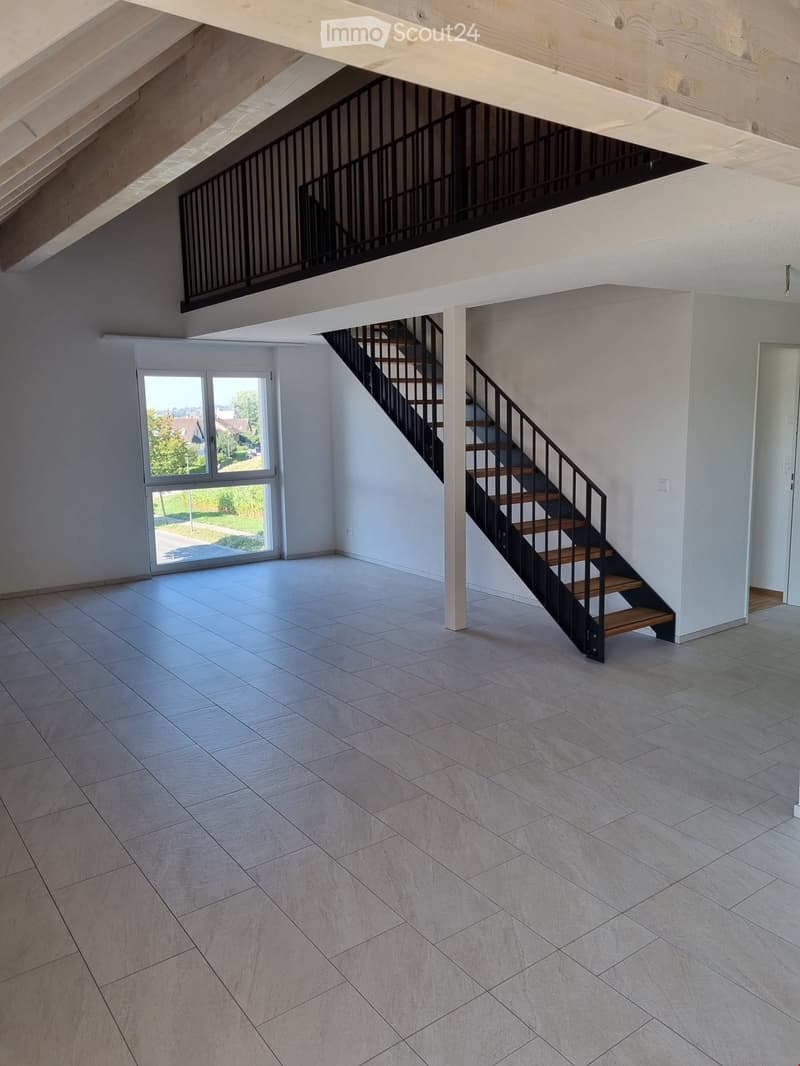 Empty room with tiled floor, staircase leading to an upper level, large window with view of greenery, and white walls.