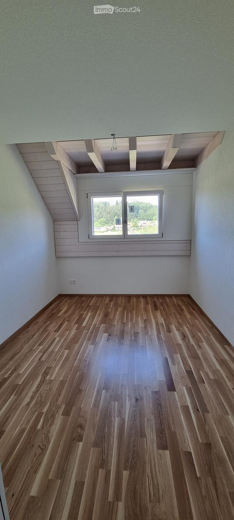 Empty room with wooden flooring, white walls, and a large window offering a view of greenery outside.