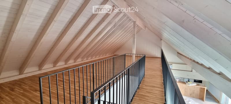 Spacious, open hallway with wooden flooring and exposed wooden beams on the ceiling. Features a modern metal railing leading to an upper level.