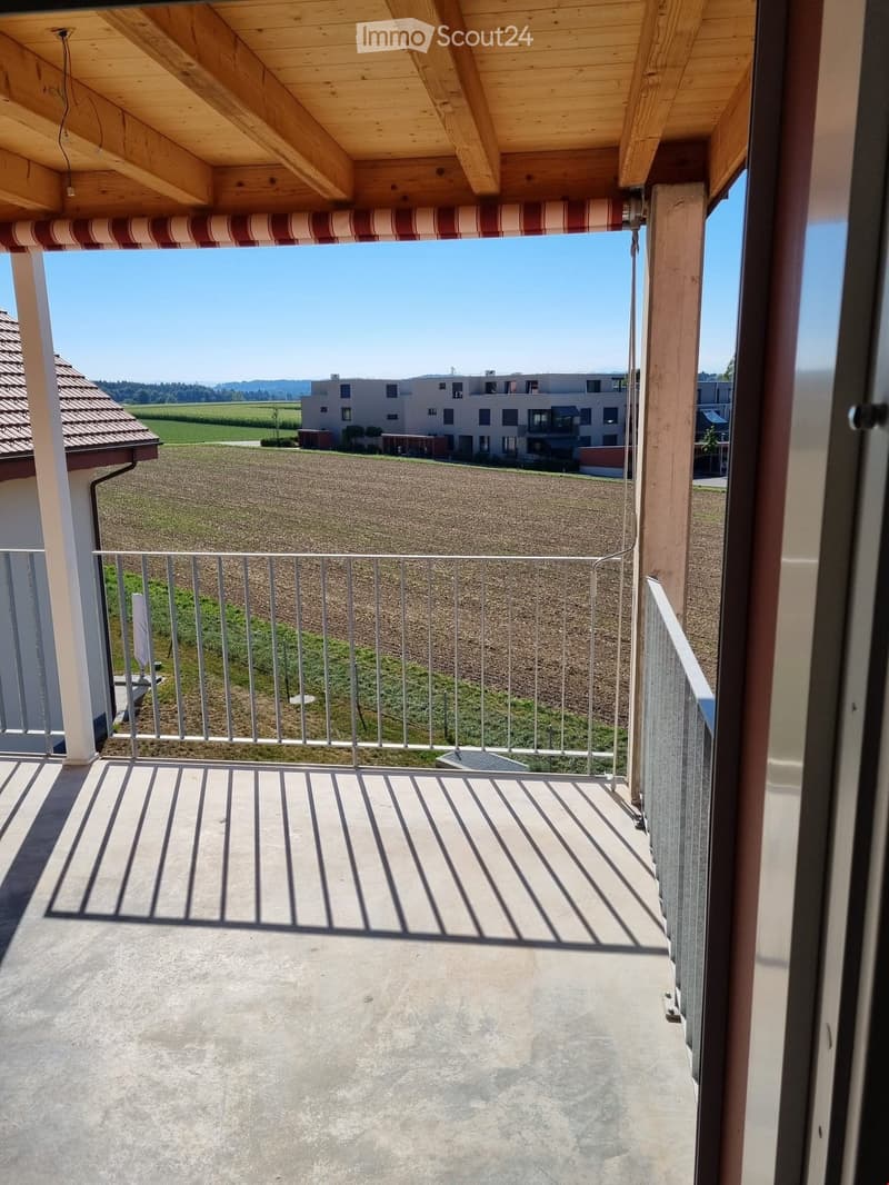Balcony with metal railing, wooden ceiling, and striped awning, overlooking a rural landscape with fields and a building in the distance.
