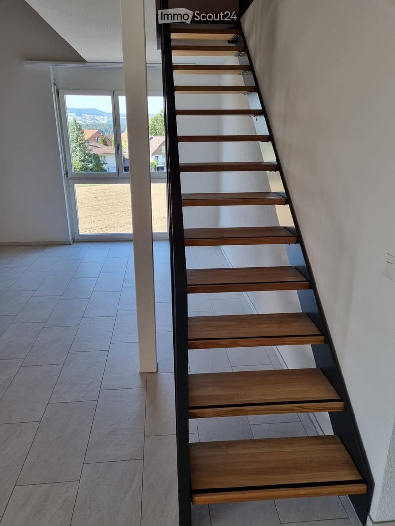 Modern staircase with wooden steps and metal railing, leading to a bright open area with large windows and a view of the landscape.