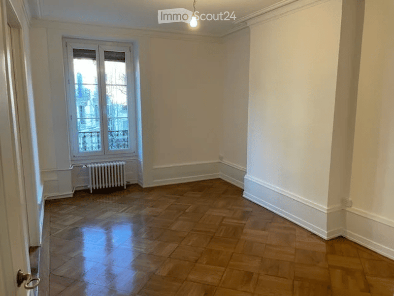 Empty room with wooden flooring, white walls, large window with view, radiator, and simple lighting fixture.