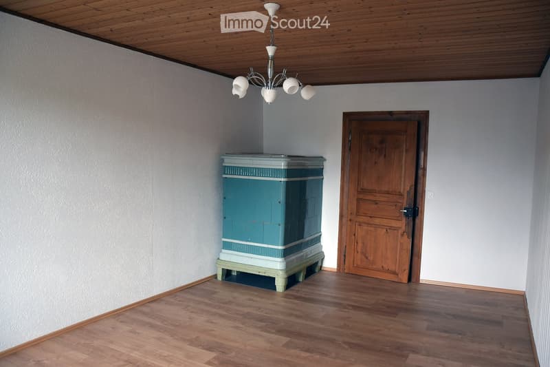 Empty room with wooden flooring, white walls, a wooden door, and a decorative stove with a chandelier above.