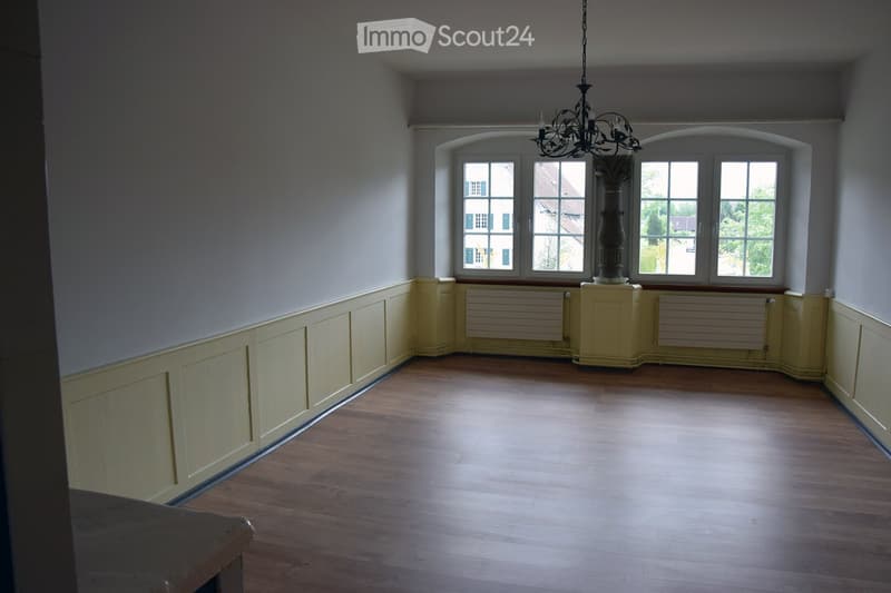 Empty room with wooden flooring, light-colored walls, and large windows with radiators beneath them. A chandelier hangs from the ceiling.