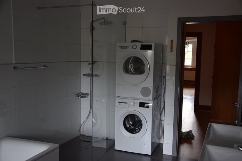 Modern bathroom with a glass-enclosed shower, a bathtub, and stacked white washing machine and dryer unit.