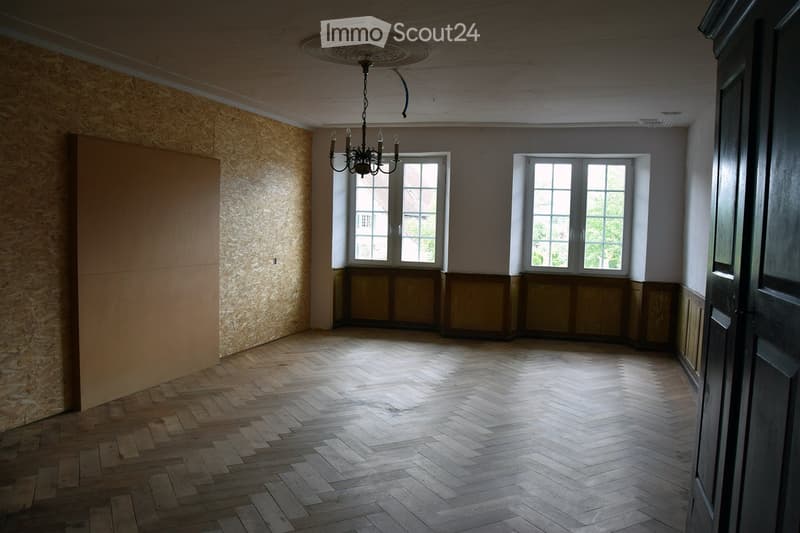 Empty room with wooden herringbone flooring, large windows, and a chandelier hanging from the ceiling.