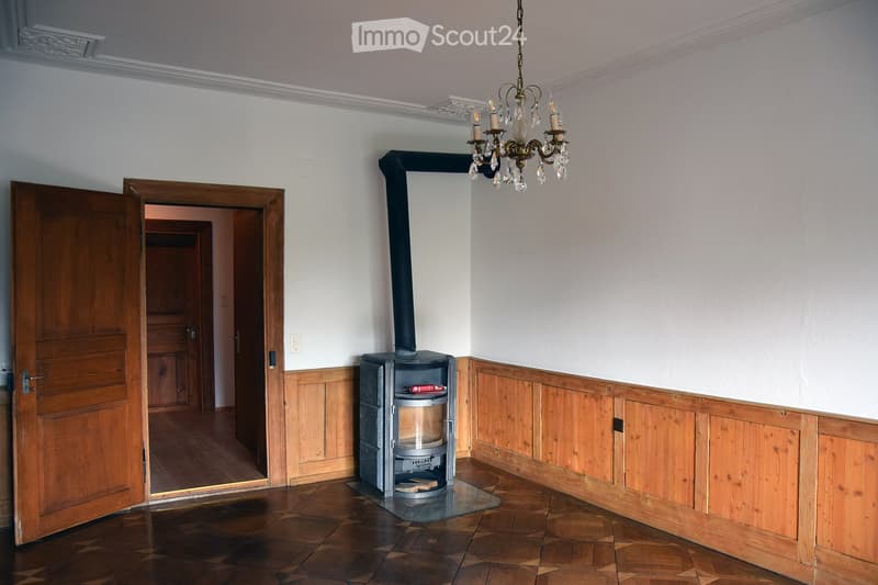 Empty room with wooden flooring, white walls, and a fireplace with a chimney. A chandelier hangs from the ceiling, and an open wooden door leads to another room.