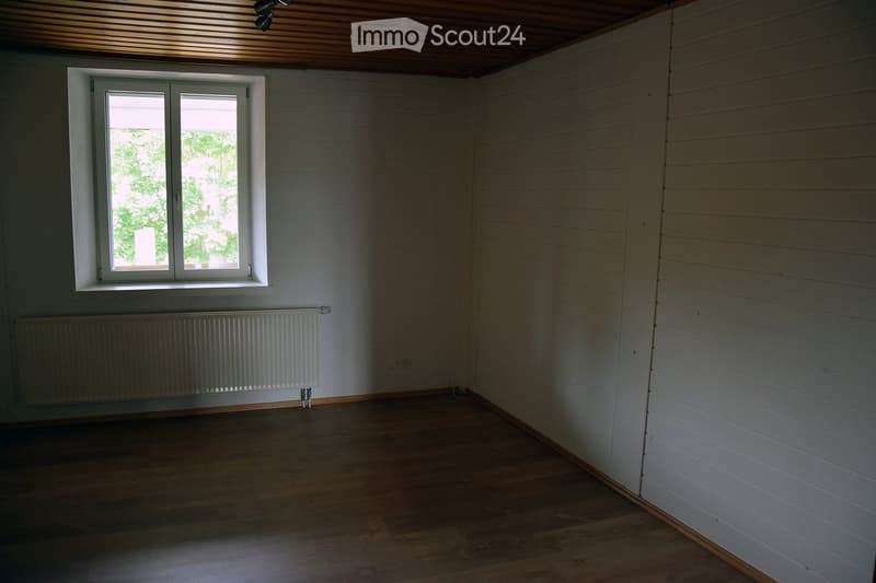 Empty room with wooden flooring, white paneled walls, a single window with white frame, and a radiator below the window.