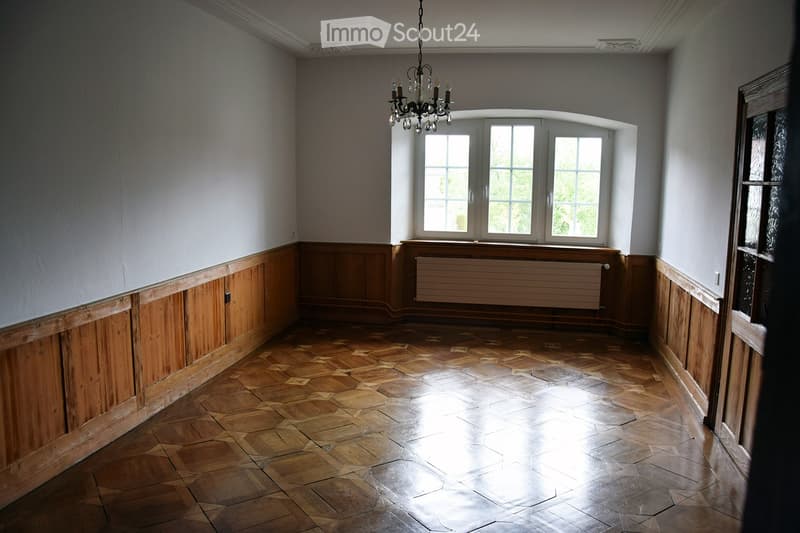 Empty room with wooden panel walls, hexagonal wooden floor, large arched window, chandelier, and radiator.