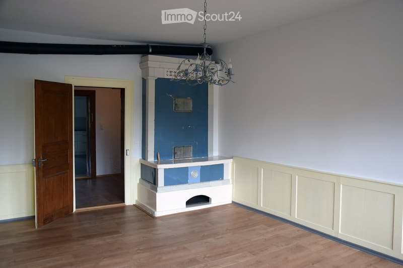 Empty room with wooden flooring, white walls, and a fireplace with a blue surround. A chandelier hangs from the ceiling, and a wooden door is open to another room.