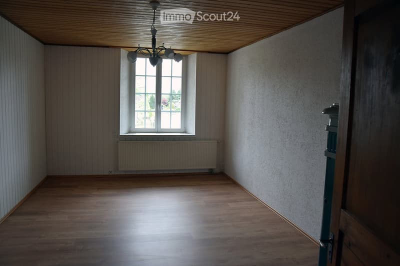 Empty room with wooden flooring, white walls, a large window with white frame, radiator below the window, and a chandelier hanging from the ceiling.