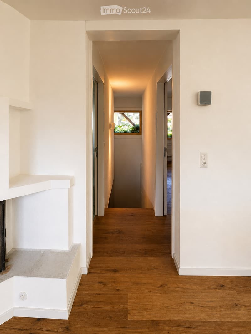 Modern hallway with wooden flooring, white walls, recessed lighting, and a small window showing greenery outside.