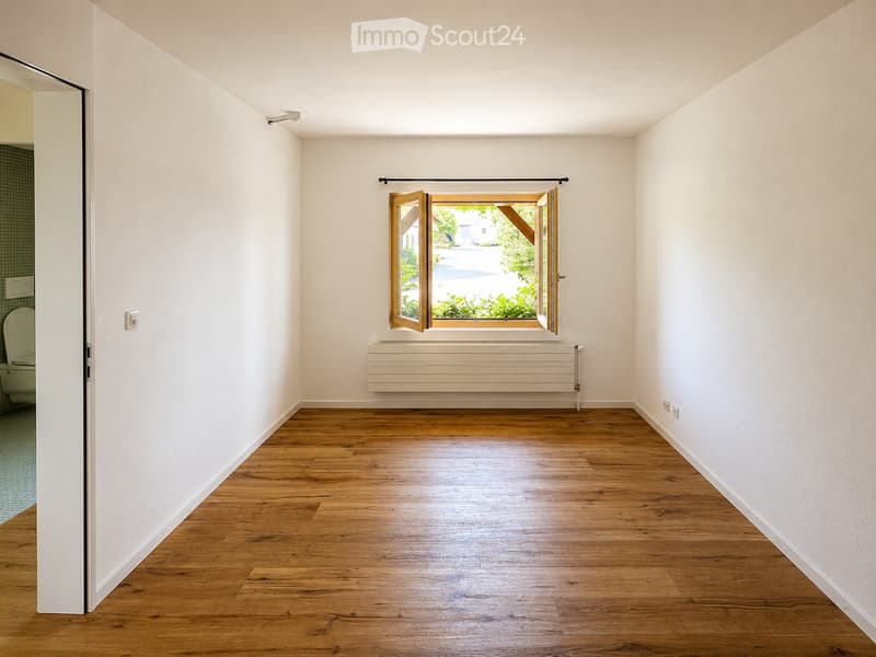 Empty room with wooden flooring, white walls, a large window with wooden frame, radiator, and a partially visible toilet in the adjacent room.