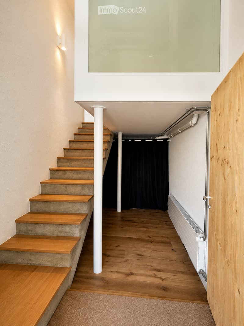 Modern hallway with wooden flooring, concrete steps leading up, white columns, and a large window at the top.
