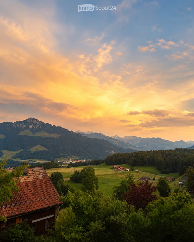 A scenic view of a rural village nestled in a lush green valley surrounded by mountains, with a wooden house in the foreground and scattered houses in the distance.