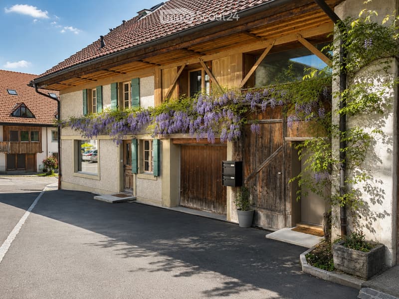 Traditional two-story building with a tiled roof, wooden shutters, and a covered wooden balcony adorned with purple wisteria flowers. The entrance features a wooden door and a small porch with a mailbox and potted plants.