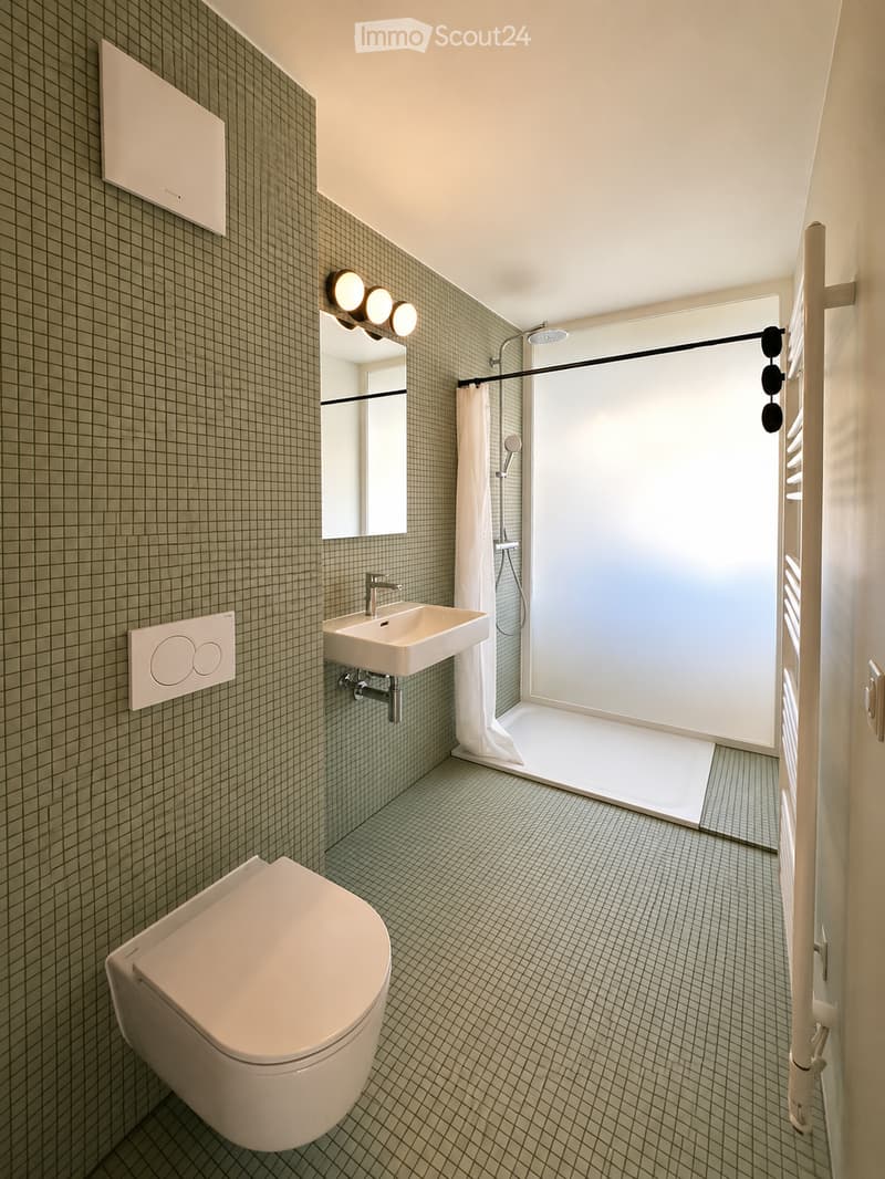 Modern bathroom with green tiled walls and floor, featuring a toilet, sink, mirror, and walk-in shower with a glass enclosure and white curtain.