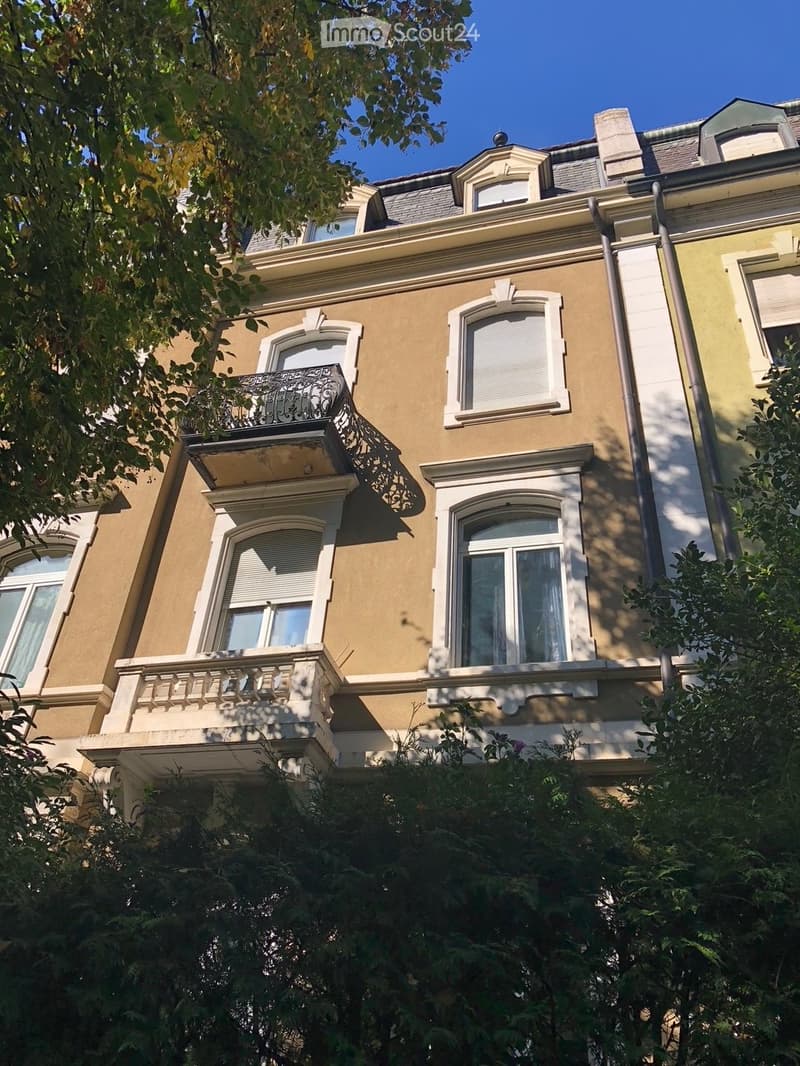 A multi-story residential building with beige facade, white window frames, ornate balconies, and decorative architectural details.