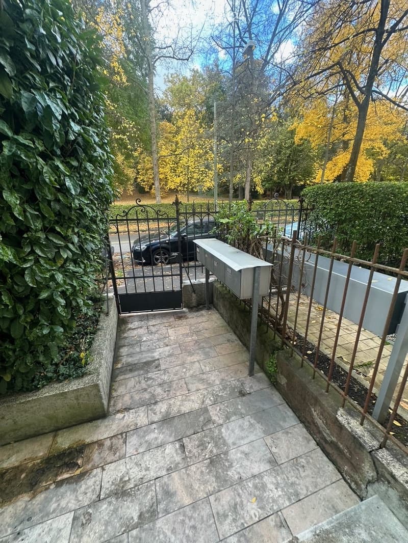 A paved pathway leads to a gated entrance with a mailbox and a parked car visible beyond the gate. The surrounding area is lined with greenery and trees.