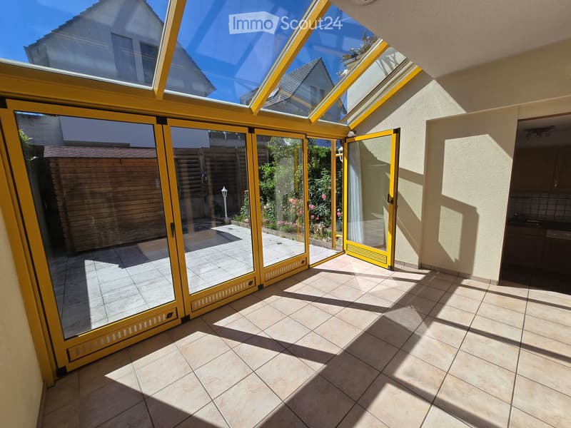 Bright sunlit balcony with large glass doors, tiled floor, and open view to a garden area.