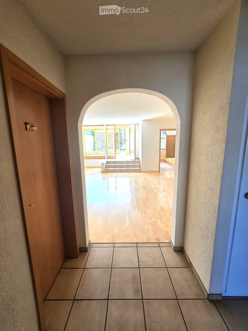 Hallway with tiled floor, wooden door on left, arched opening leading to another room with wooden flooring and large windows.