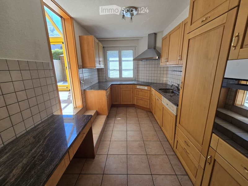 Modern kitchen with light wooden cabinets, tiled walls, and a dark countertop. Features a sink, faucet, and range hood.