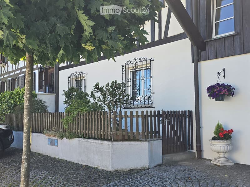 Two-story house with white walls, dark wooden trim, decorative wrought iron window grilles, and a wooden fence with small plants and trees in front.