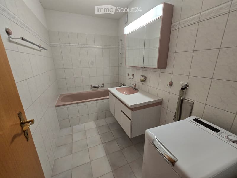 Bathroom with a bathtub, sink, mirror, and cabinet. The walls and floor are tiled, and a washing machine is visible in the corner.