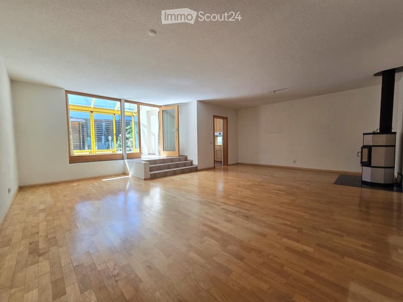 Empty room with wooden flooring, large windows, and a small staircase leading outside. A wood-burning stove is present in the corner.