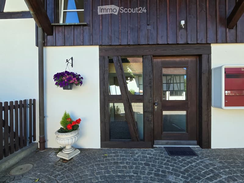 Two-story building with a wooden facade, a white lower section, a wooden front door with glass panels, a hanging flower basket, a decorative urn with flowers, and a mailbox.