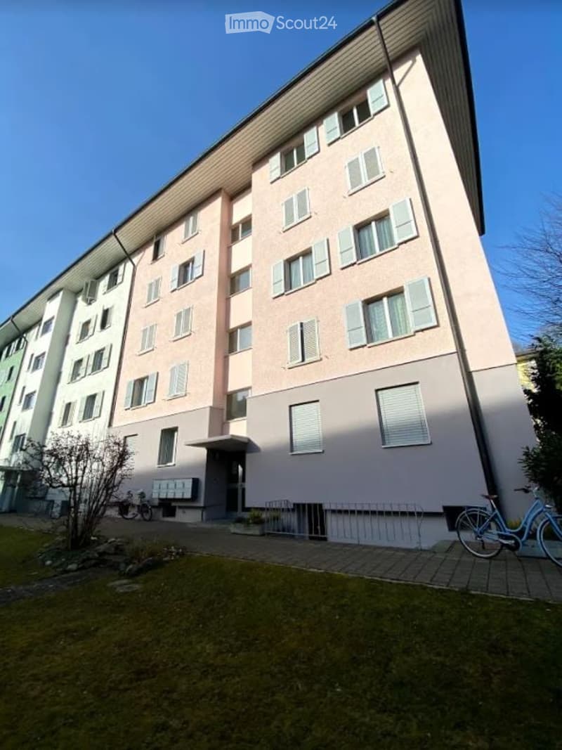 Multi-story residential building with light pink facade, white shutters, and a small entrance area with a bicycle rack.