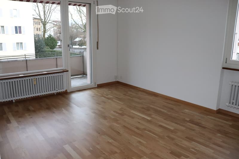 Empty room with light wooden flooring, white walls, and large windows with blinds. Radiators are visible along the walls, and there is a sliding glass door leading to a balcony area.