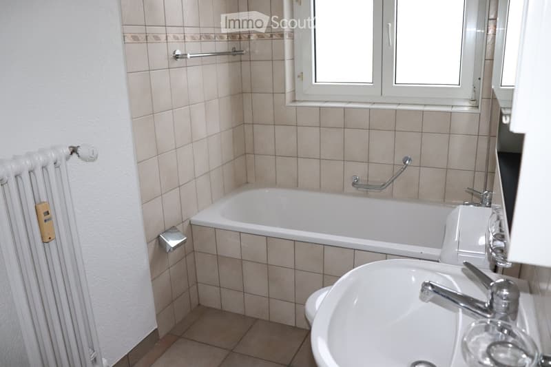 Bathroom with a bathtub, sink, and radiator. The walls are tiled with beige tiles and a decorative border. A window is above the bathtub, and a handrail is attached to the wall next to the bathtub.