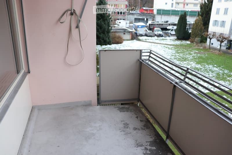 Empty balcony with metal railing, concrete floor, and a view of a snowy outdoor area with buildings and vehicles.