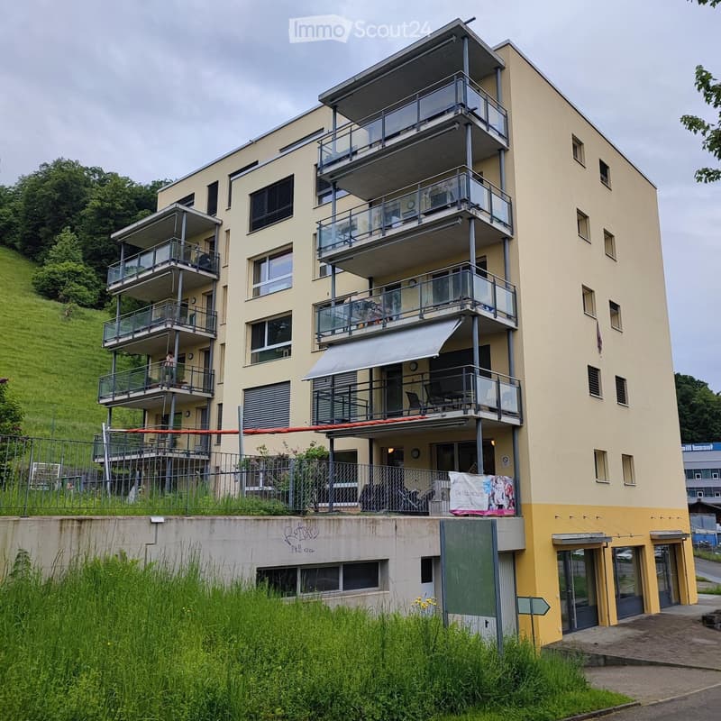 Modern multi-story residential building with balconies, large windows, and a raised ground floor. The building has a yellow facade and is surrounded by greenery.
